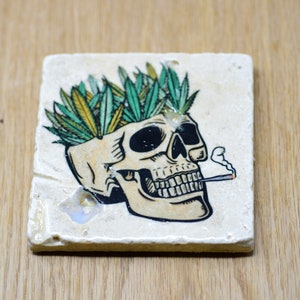 May include: A white ceramic coaster with a skull design. The skull is wearing a crown of green marijuana leaves and is smoking a cigarette.