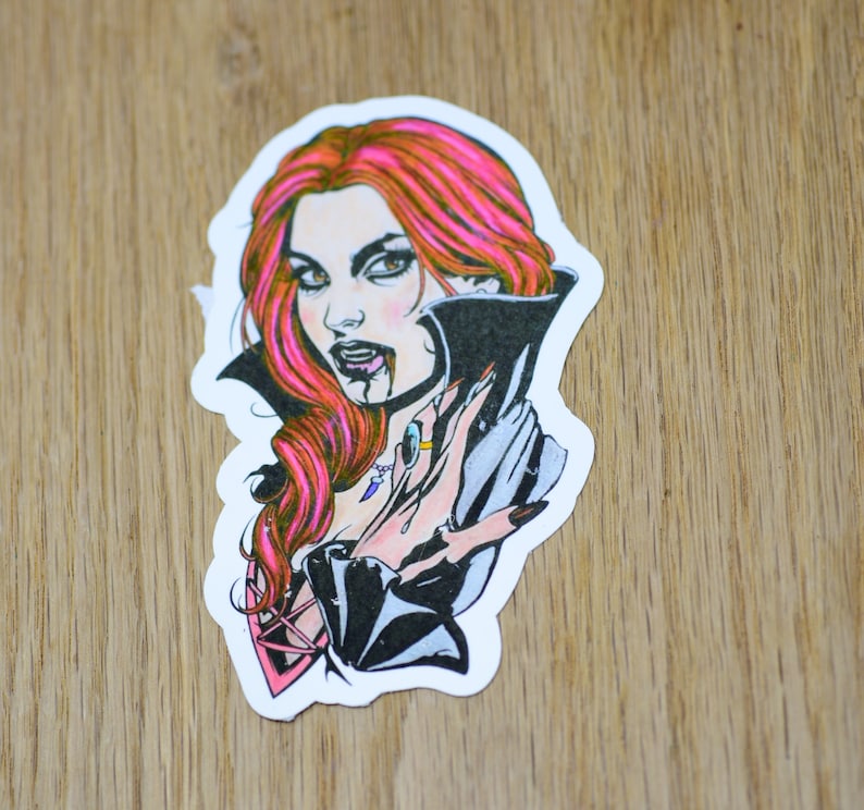 Puede incluir: Una pegatina con una ilustraci&oacute;n colorida de una mujer vampiro con cabello rojo, usando una capa negra y sosteniendo una mano frente a su cara. La mujer tiene colmillos y le gotea sangre de la boca.