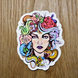 May include: A colorful sticker featuring a woman with snakes in her hair. The woman has a pink rose in her hair and her eyes are closed. The snakes are green, blue, and yellow. The background is white.