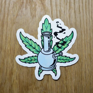 May include: A white glass bong with a green marijuana leaf design. The bong has a round base and a long, thin neck. There is a black smoke design coming out of the top of the bong.