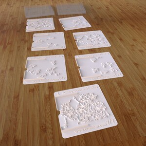 May include: Set of six white plastic cookie cutters with various floral designs. The cutters are square shaped and have a raised design. The cutters are labeled "Morning Glory and Tea Rose" and include the website address "www.bromblan.com".