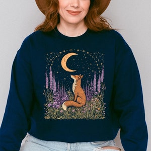 Op de afbeelding: Een donkerblauw sweatshirt met een afbeelding van een vos die opkijkt naar een halve maan in een veld met paarse bloemen. De maan is geel en de sterren zijn goud. De tekst "Shroom Queens" staat op het sweatshirt gedrukt.