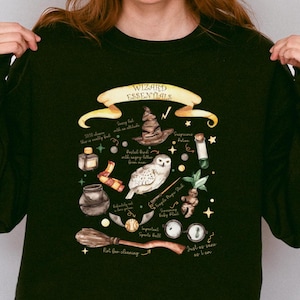 Könnte beinhalten: Ein schwarzes Sweatshirt mit einem grafischen Druck von Zauberer-Essentials, darunter ein Hut, eine Eule, Zaubertrankflaschen und ein Zauberstab. Der Text "Wizard Essentials" ist auf dem Grafikdruck zu sehen.