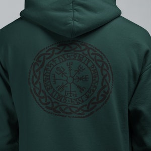 Könnte beinhalten: Ein dunkelgrüner Kapuzenpullover mit einem schwarzen und weißen Kompass-Design auf der Rückseite. Der Kompass ist von einem Kreis aus Knoten und Runen umgeben.