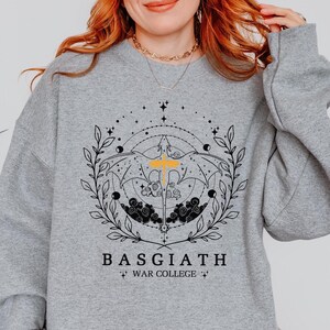 Könnte beinhalten: Grauer Pullover mit einem schwarzen und gelben Grafikdesign, das eine himmlische Szene mit einem geflügelten Wesen, Sternen und dem Text "BASGIATH WAR COLLEGE" zeigt.