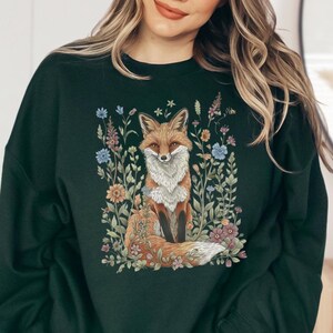 Könnte beinhalten: Ein dunkelgrüner Pullover mit einem Fuchs- und Blumenmuster. Der Fuchs ist orange und weiß mit einem buschigen Schwanz. Die Blumen sind in den Farben Rosa, Blau und Gelb gehalten.