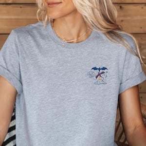 Könnte beinhalten: Ein hellgraues T-Shirt mit hochgekrempelten Ärmeln. Das Shirt hat ein kleines gesticktes Cartoon-Design eines Charakters unter einem Regenschirm. Ein Strohhut mit schwarzem Band liegt auf dem Schoß.