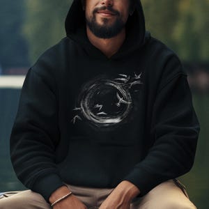 Może przedstawiać: Czarna bluza z kapturem z białym okrągłym wzorem i sylwetkami ptaków. Bluza ma przednią kieszeń i kaptur ze sznurkiem. Osoba na zdjęciu ma na sobie srebrną bransoletkę i spodnie khaki.