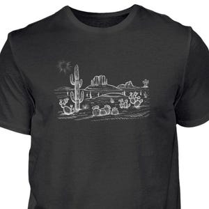 Könnte beinhalten: Schwarzes T-Shirt mit einer weißen Skizze einer Wüstenlandschaft. Das Design zeigt Kakteen, eine Sonne und eine Bergkette. Die Grafik ist mittig auf der Vorderseite platziert und bietet eine lässige und künstlerische Ästhetik.