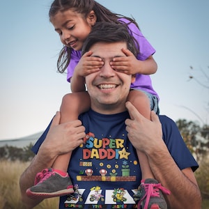 Op de afbeelding: Een marineblauw t-shirt met de tekst "SUPER DADDY" en een kleurrijke videogame-afbeelding. Een kind zit op de schouders van de man en bedekt zijn ogen. Het kind draagt een paars shirt en een spijkerbroek.