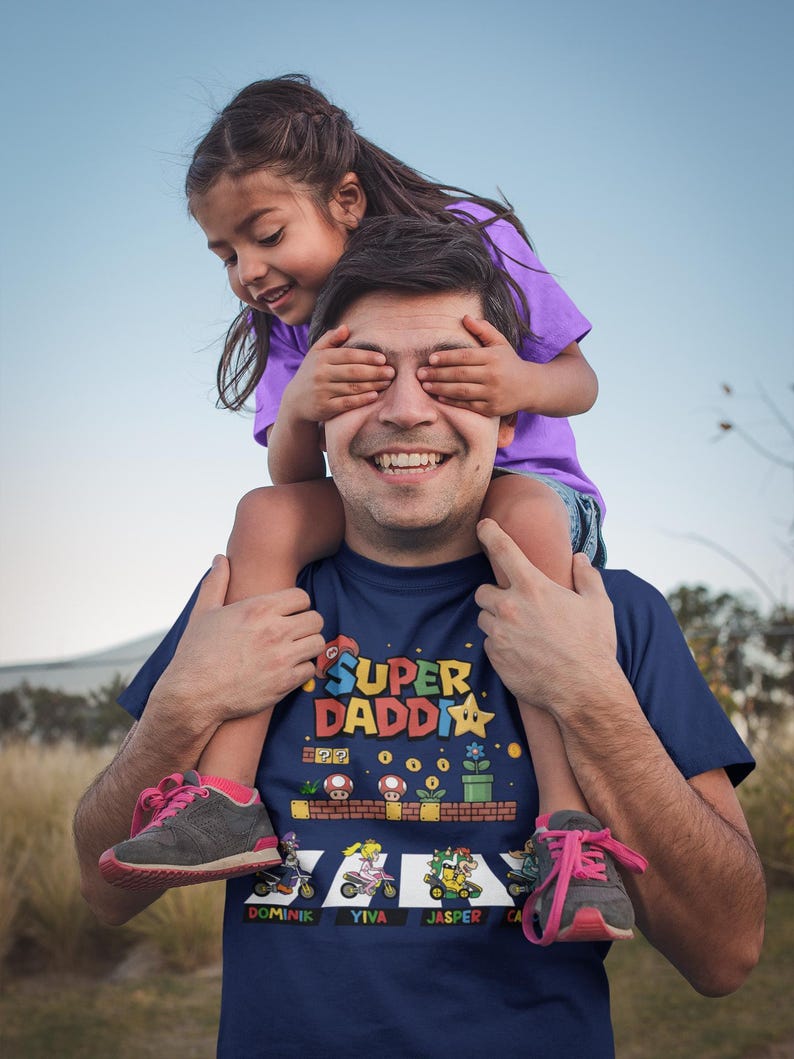 Op de afbeelding: Een marineblauw t-shirt met de tekst "SUPER DADDY" en een kleurrijke videogame-afbeelding. Een kind zit op de schouders van de man en bedekt zijn ogen. Het kind draagt een paars shirt en een spijkerbroek.