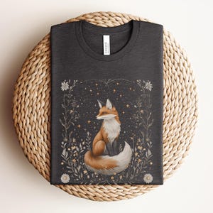 May include: A dark grey t-shirt featuring a graphic print of a fox sitting in a field of flowers and stars. The fox is orange and white with a bushy tail.