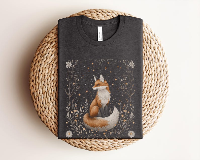 May include: A dark grey t-shirt featuring a graphic print of a fox sitting in a field of flowers and stars. The fox is orange and white with a bushy tail.