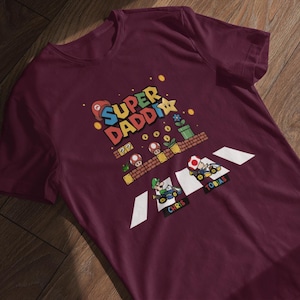 Op de afbeelding: Bordeauxrood t-shirt met een kleurrijke afbeelding met de tekst "Super Daddi" en personages uit een videogame. Het ontwerp bevat een gepixeld landschap met paddenstoelen, bloemen en twee go-karts met de namen "Chris" en "Tobias".