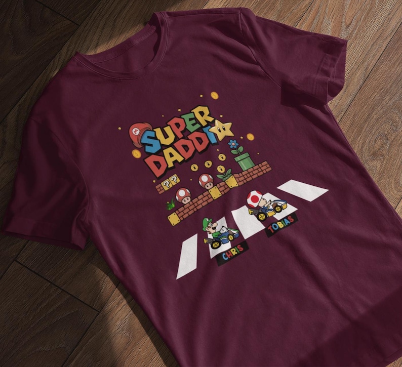 Op de afbeelding: Bordeauxrood t-shirt met een kleurrijke afbeelding met de tekst "Super Daddi" en personages uit een videogame. Het ontwerp bevat een gepixeld landschap met paddenstoelen, bloemen en twee go-karts met de namen "Chris" en "Tobias".