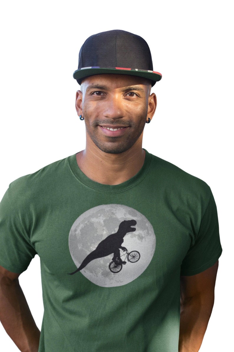 May include: A man wearing a green t-shirt with a black silhouette of a dinosaur riding a bicycle on the moon. He is also wearing a black baseball cap with a red, white, and blue stripe.