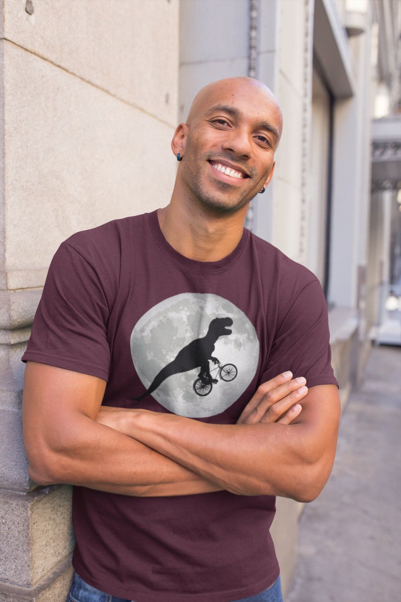 May include: A maroon t-shirt with a graphic of a dinosaur riding a bicycle in front of a full moon.