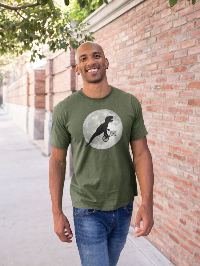 May include: A man wearing a green t-shirt with a black silhouette of a dinosaur riding a bicycle on the moon.