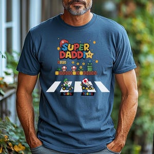 Op de afbeelding: Marineblauw T-shirt met een kleurrijke afbeelding met de tekst "Super Daddi" en een ontwerp met een videogame-thema. Het ontwerp bevat personages in go-karts, paddenstoelen en een ster. De namen "Chris" en "Tobias" zijn ook zichtbaar.