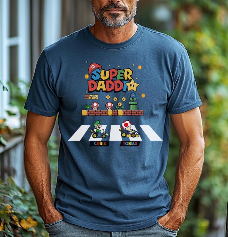 Op de afbeelding: Marineblauw T-shirt met een kleurrijke afbeelding met de tekst "Super Daddi" en een ontwerp met een videogame-thema. Het ontwerp bevat personages in go-karts, paddenstoelen en een ster. De namen "Chris" en "Tobias" zijn ook zichtbaar.