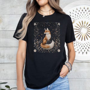 May include: Black t-shirt featuring a graphic of a fox sitting in a field of flowers and stars. The fox is orange and white with a bushy tail.