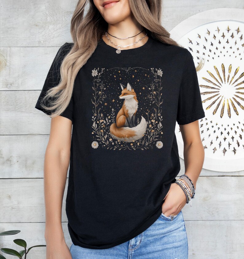 May include: Black t-shirt featuring a graphic of a fox sitting in a field of flowers and stars. The fox is orange and white with a bushy tail.