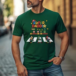 Op de afbeelding: Een donkergroen t-shirt met de tekst "Super Daddi" en een Mario-thema-afbeelding. De afbeelding toont personages in go-karts met de namen "Anna", "Patrick" en "Thomas" eronder. Het shirt wordt door een persoon gedragen.
