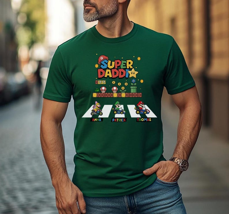 Op de afbeelding: Een donkergroen t-shirt met de tekst "Super Daddi" en een Mario-thema-afbeelding. De afbeelding toont personages in go-karts met de namen "Anna", "Patrick" en "Thomas" eronder. Het shirt wordt door een persoon gedragen.