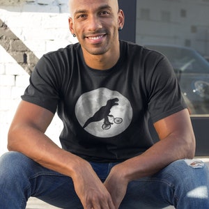 May include: A black t-shirt with a white circle graphic of a dinosaur riding a bicycle on the moon.