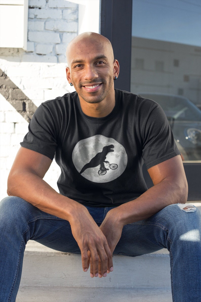 May include: A black t-shirt with a white circle graphic of a dinosaur riding a bicycle on the moon.