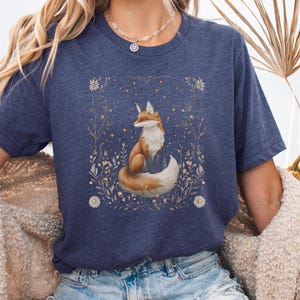 May include: A navy blue t-shirt featuring a graphic of a fox sitting in a field of flowers. The fox is orange and white with a bushy tail. The flowers are white and yellow.