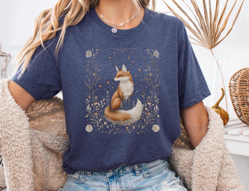 May include: A navy blue t-shirt featuring a graphic of a fox sitting in a field of flowers. The fox is orange and white with a bushy tail. The flowers are white and yellow.