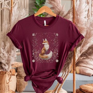 May include: A maroon t-shirt featuring a graphic of a fox sitting in a field of flowers and stars. The fox is orange and white with a bushy tail.