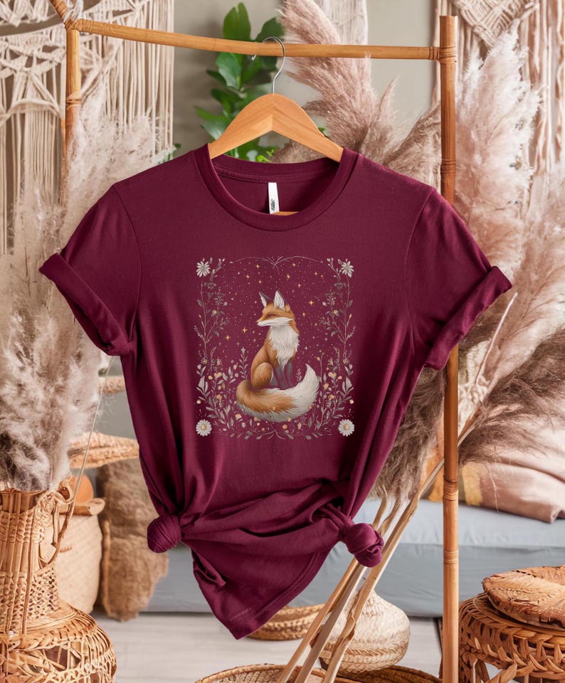 May include: A maroon t-shirt featuring a graphic of a fox sitting in a field of flowers and stars. The fox is orange and white with a bushy tail.