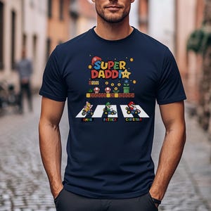 Op de afbeelding: Marineblauw T-shirt met een kleurrijke afbeelding met de tekst "Super Daddy" en een videogame-thema ontwerp. Het ontwerp bevat personages in go-karts met namen eronder. Een leuk en speels ontwerp.