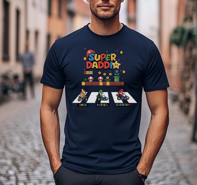 Op de afbeelding: Marineblauw T-shirt met een kleurrijke afbeelding met de tekst "Super Daddy" en een videogame-thema ontwerp. Het ontwerp bevat personages in go-karts met namen eronder. Een leuk en speels ontwerp.
