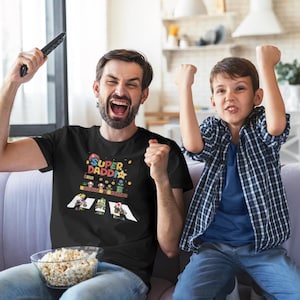 Op de afbeelding: Zwart t-shirt met een kleurrijke Super Daddio-afbeelding, met een videogame-thema met personages en tekst. Het shirt wordt gedragen door een man die een afstandsbediening vasthoudt en viert met een kind.
