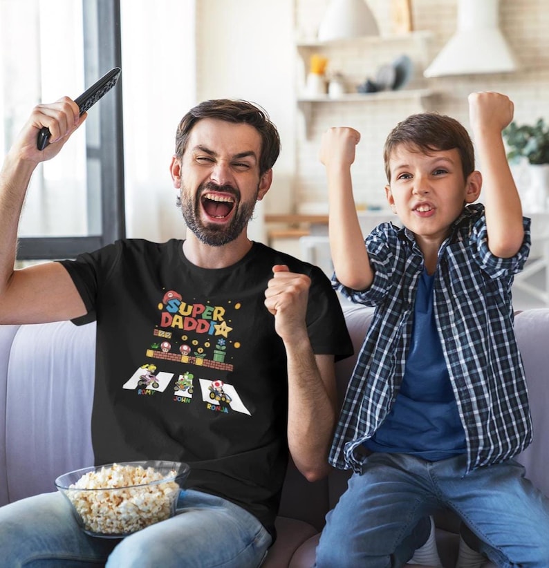 Op de afbeelding: Zwart t-shirt met een kleurrijke Super Daddio-afbeelding, met een videogame-thema met personages en tekst. Het shirt wordt gedragen door een man die een afstandsbediening vasthoudt en viert met een kind.
