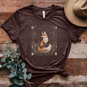 May include: A brown t-shirt featuring a graphic of a fox sitting in a field of flowers. The fox is orange and white with a bushy tail. The flowers are white and yellow.