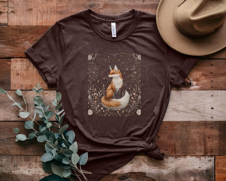 May include: A brown t-shirt featuring a graphic of a fox sitting in a field of flowers. The fox is orange and white with a bushy tail. The flowers are white and yellow.