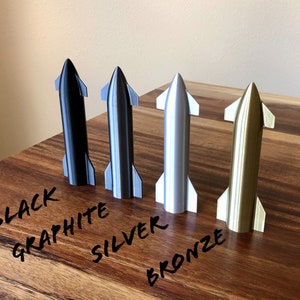 May include: Four miniature rocket models in black, graphite, silver, and bronze. The rockets are displayed on a wooden surface, with handwritten labels identifying each color. The rockets have a sleek, aerodynamic design.