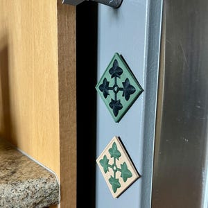 May include: A silver missile-shaped magnet and two diamond-shaped magnets with a green and black design. The magnets are attached to a gray refrigerator door. The diamond magnets have a four-leaf clover design.
