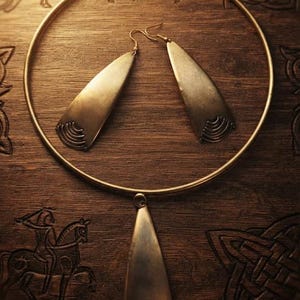 Peut inclure: Un collier torque en métal doré avec un pendentif triangulaire central et une paire de boucles d'oreilles pendantes triangulaires assorties. Chaque pièce présente une surface lisse avec des lignes gravées courbes à la base.