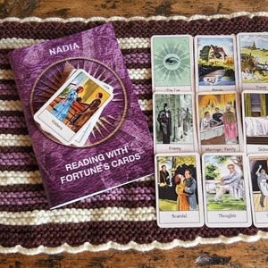 Op de afbeelding: Een paars boek getiteld Reading with Fortune's Cards door Nadia, met een cirkelvormige afbeelding en een kleine kaart op de omslag. Negen rechthoekige waarzegkaarten zijn in een raster naast het boek op een gestreepte gebreide mat geplaatst.