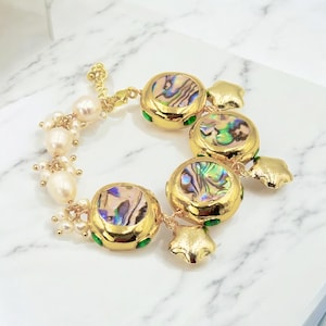 Pode incluir: Pulseira dourada com detalhes iridescentes de concha de abalone em molduras redondas. A pulseira inclui fios de pérolas e pingentes em forma de estrela. O design incorpora elementos verdes e dourados, criando uma joia luxuosa e atraente.
