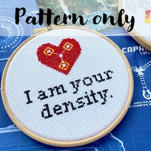 May include: Cross stitch pattern featuring a red heart with a yellow symbol inside, and the text "I am your density." in black lettering. The pattern is displayed in a wooden embroidery hoop.