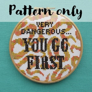 Könnte beinhalten: Ein Kreuzstich-Stickrahmen mit weißem Hintergrund und einem bunten Muster. Der Text "VERY DANGEROUS... YOU GO FIRST" ist in schwarzem Garn gestickt.