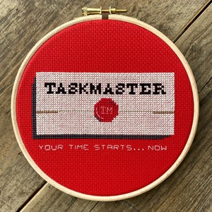 May include: A red embroidery hoop with a white envelope that says "Taskmaster" with a red wax seal that says "TM". The text below the envelope says "Your time starts... now".