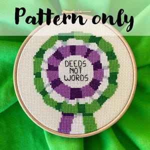 May include: Cross-stitch embroidery hoop with a green, white, and purple design. The text "DEEDS NOT WORDS" is centered within the design. The words "Pattern only" are at the top of the image.