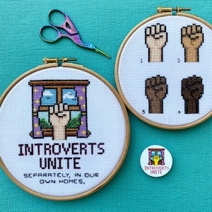 May include: Embroidered cross-stitch hoops with various designs. One hoop displays a fist in a window with the text "Introverts Unite." Another shows four fists in different skin tones. A pair of decorative scissors and a button are also visible.